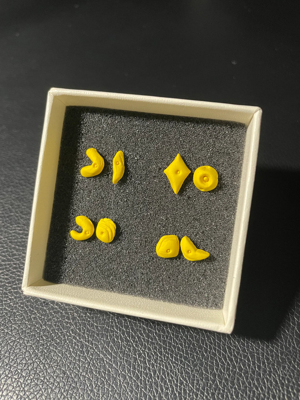 Yellow earrings in the shape of climbing holds in a square display case on a dark textured surface